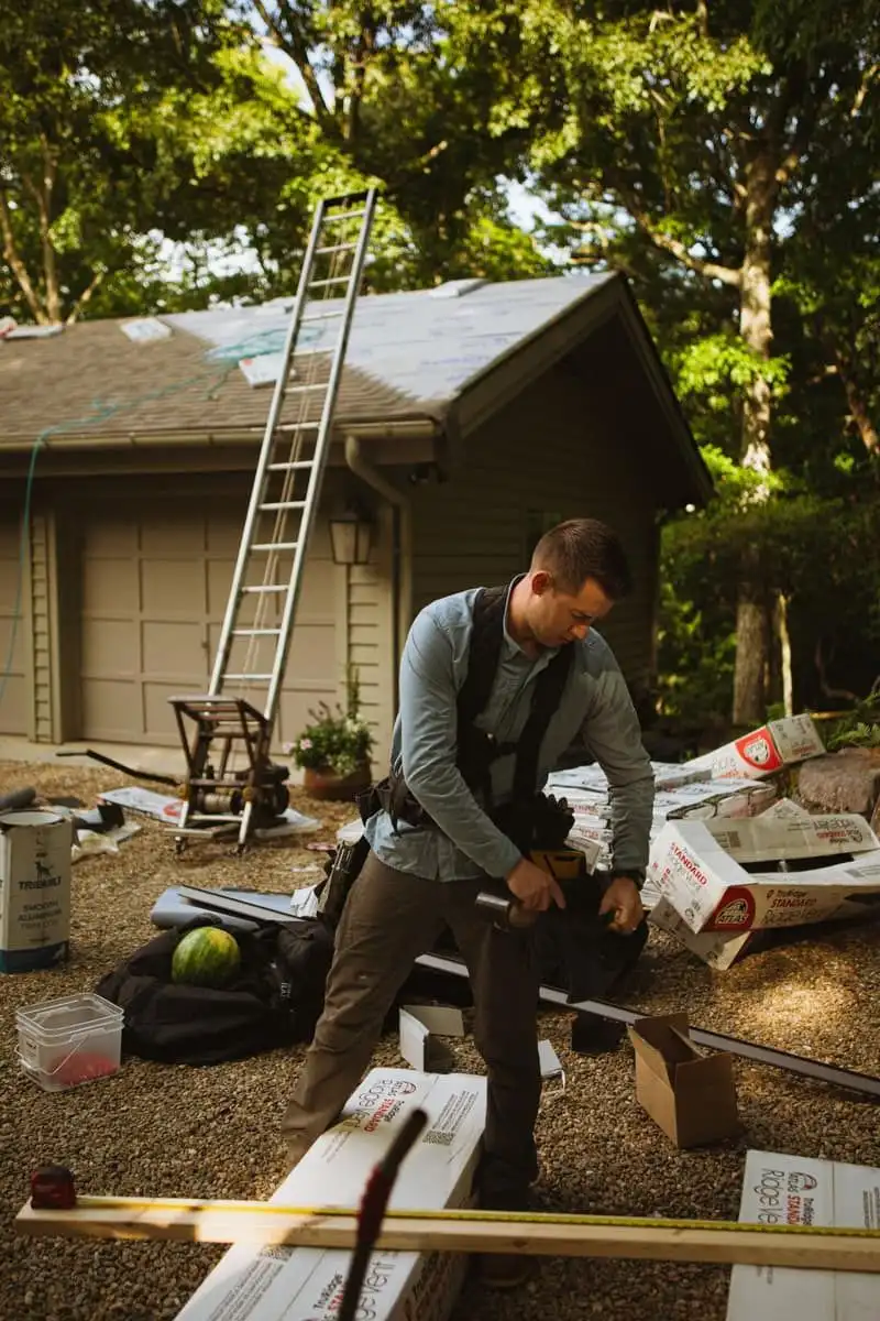 A roofer in a cap and tool belt, seen from the side, working on a residential roof